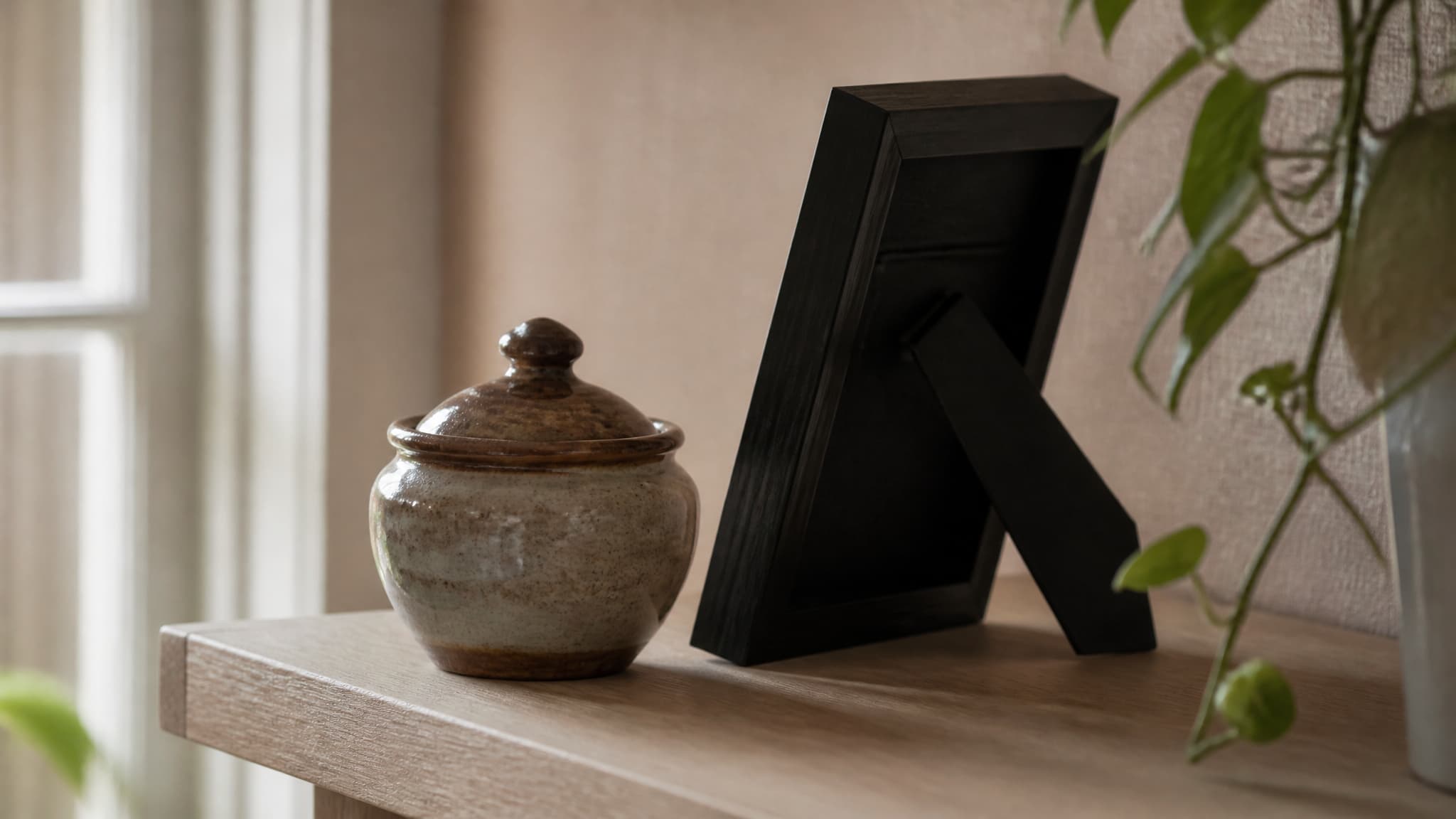 A small glazed ceramic urn with a fitted lid standing on a pale wooden shelf beside a framed photograph turned slightly away, soft window light from the left, a trailing houseplant at the edge of the frame
