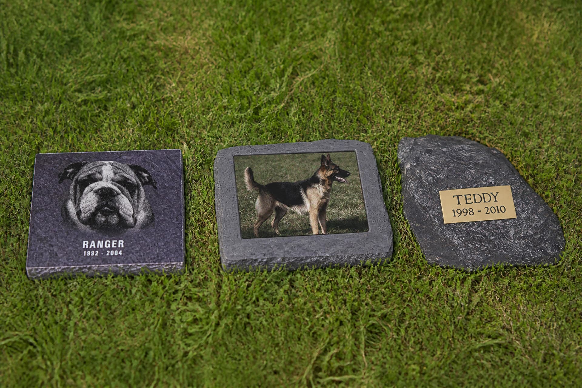 Three indoor pet memorial pieces arranged on a pale wooden sideboard in soft window light, a standing engraved plaque, a small framed photo memorial and a low stone keepsake, each with a smooth finished face