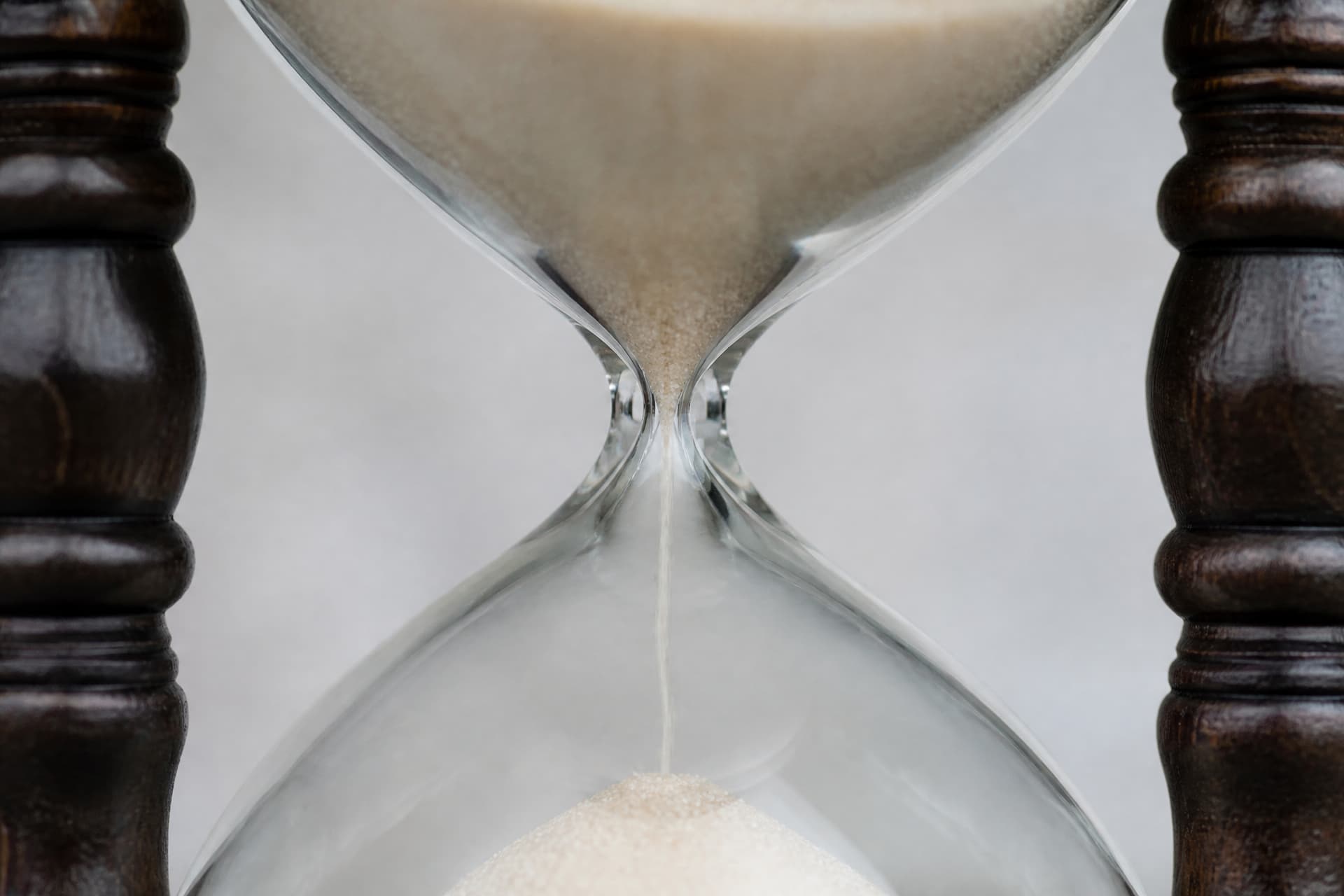 A close view of a sealed glass hourglass in a dark turned wooden frame on a pale surface, the narrow waist between the two chambers clearly visible with fine pale grains part-way through