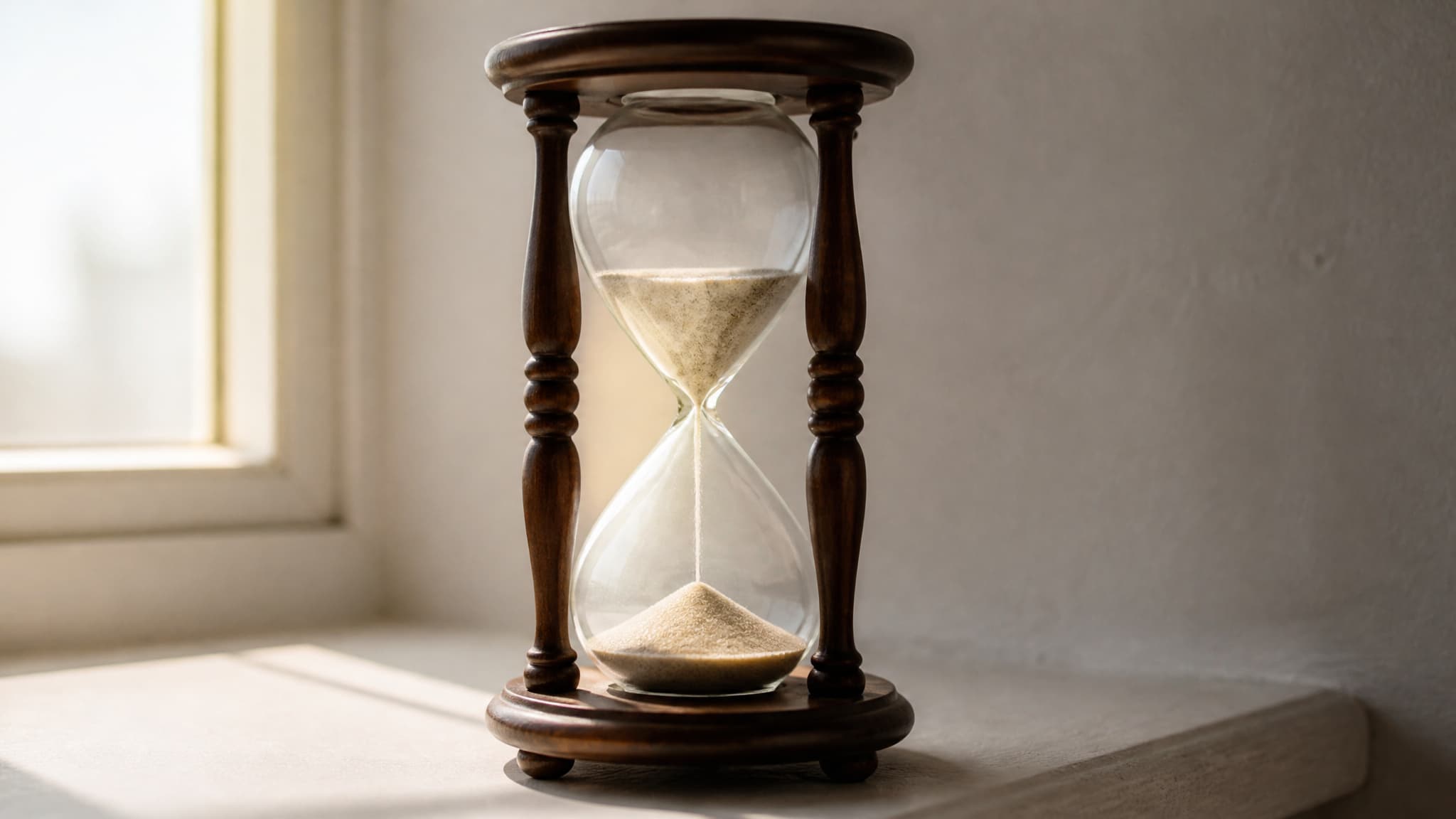 A sealed glass hourglass in a turned wooden frame standing on a pale windowsill in soft morning light, fine pale sand mid-fall between the two chambers