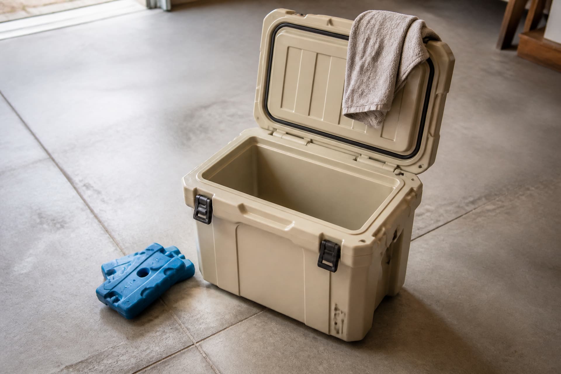A hard-sided cooler box standing open and empty on a clean garage floor in flat daylight, two blue freezer packs resting beside it, a folded towel on the lid