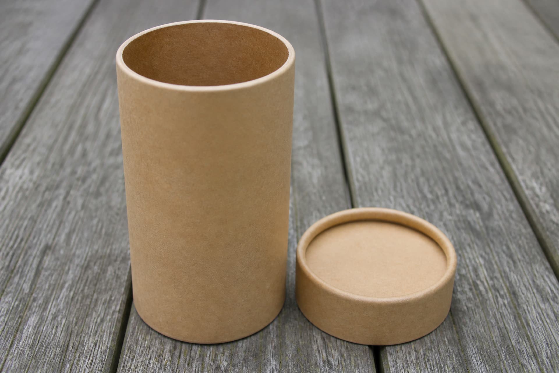 A plain kraft-brown cardboard cylinder about ten inches tall standing on weathered timber in flat daylight, its separate push-on lid set beside it, the open mouth showing a smooth uncoated inner wall