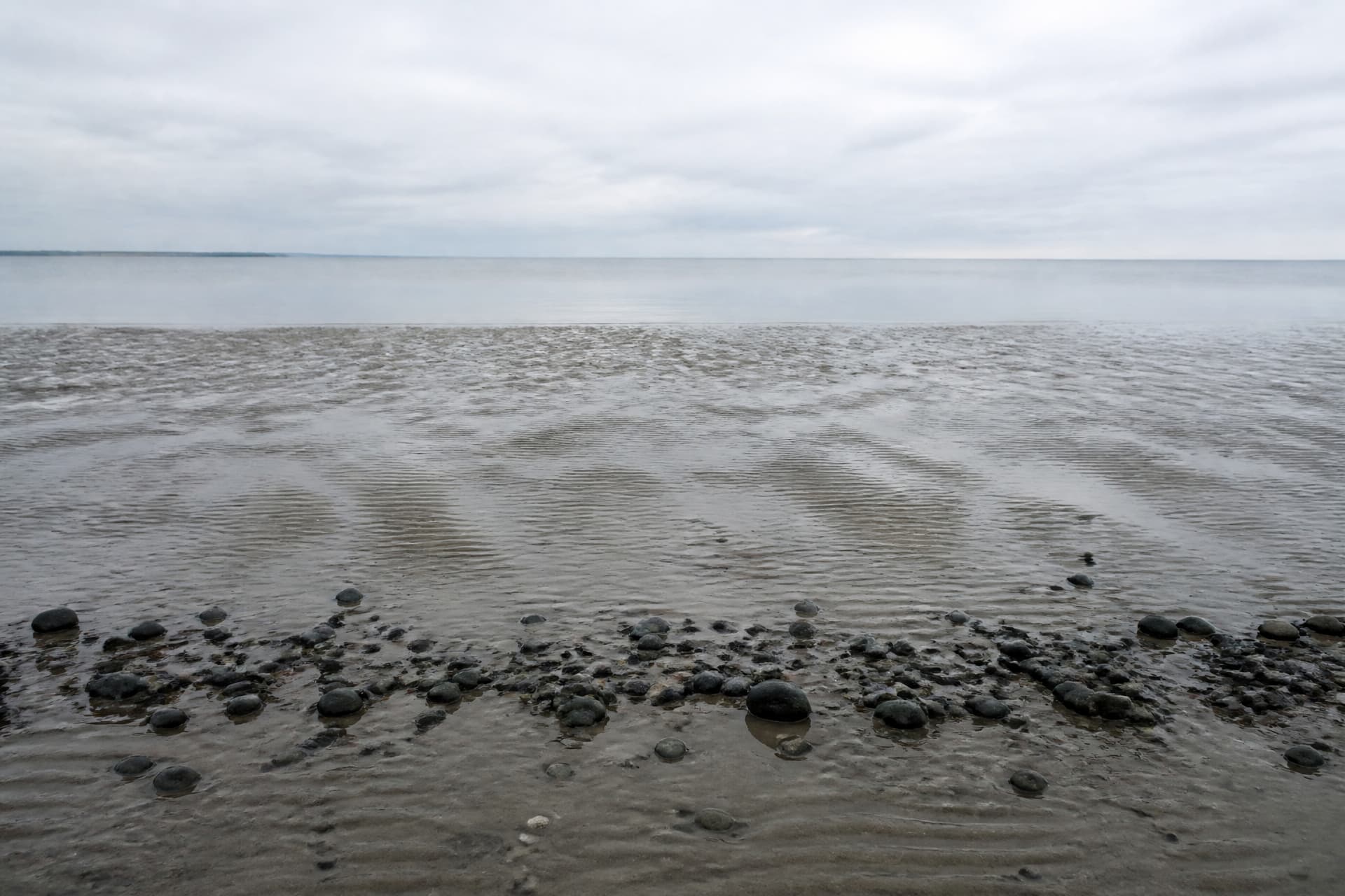 A shoreline at low tide in flat overcast light, wet sand reflecting a pale sky, gentle water at the far edge and a line of dark pebbles in the foreground, no people or objects
