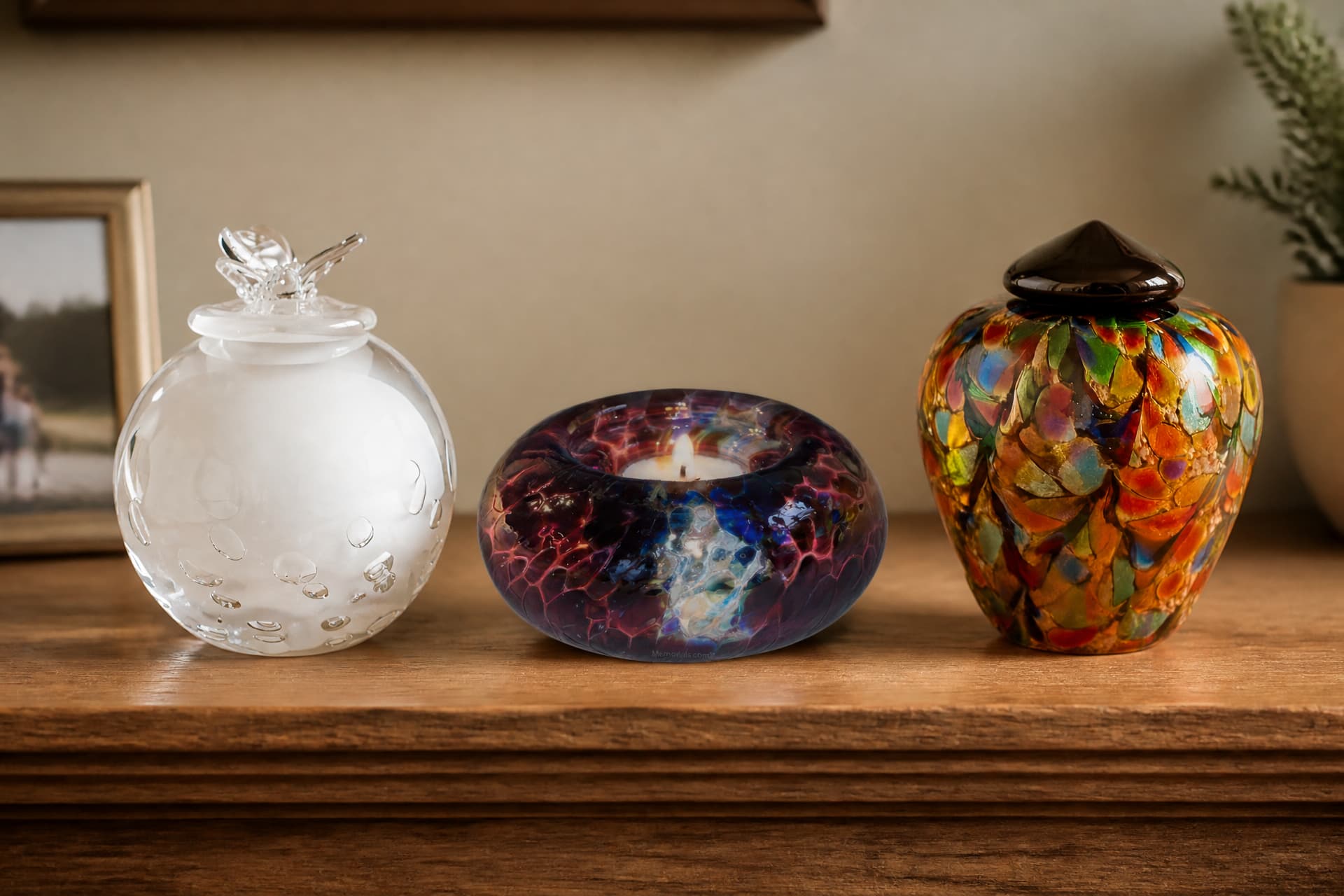 Three glass pet urns on a pale wooden shelf in soft window light, one rounded amber vessel, one tall clear cylinder and one swirled blue-green form, each with a fitted base and a smooth polished surface