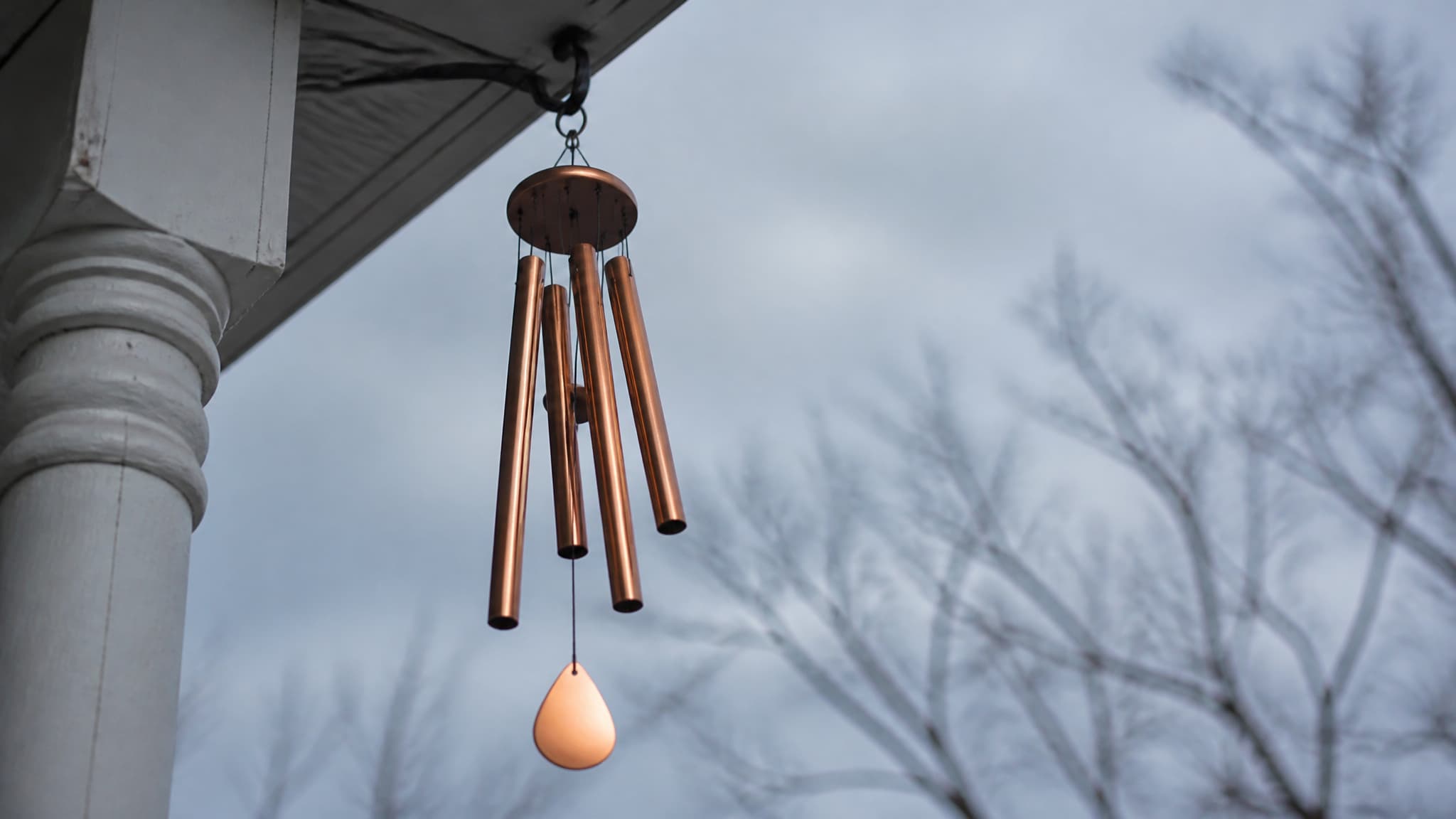 A set of five brushed copper wind chime tubes and a teardrop wind catcher hanging from the eave of a porch against a soft gray morning sky, the tubes caught mid-sway with a bare maple branch behind them