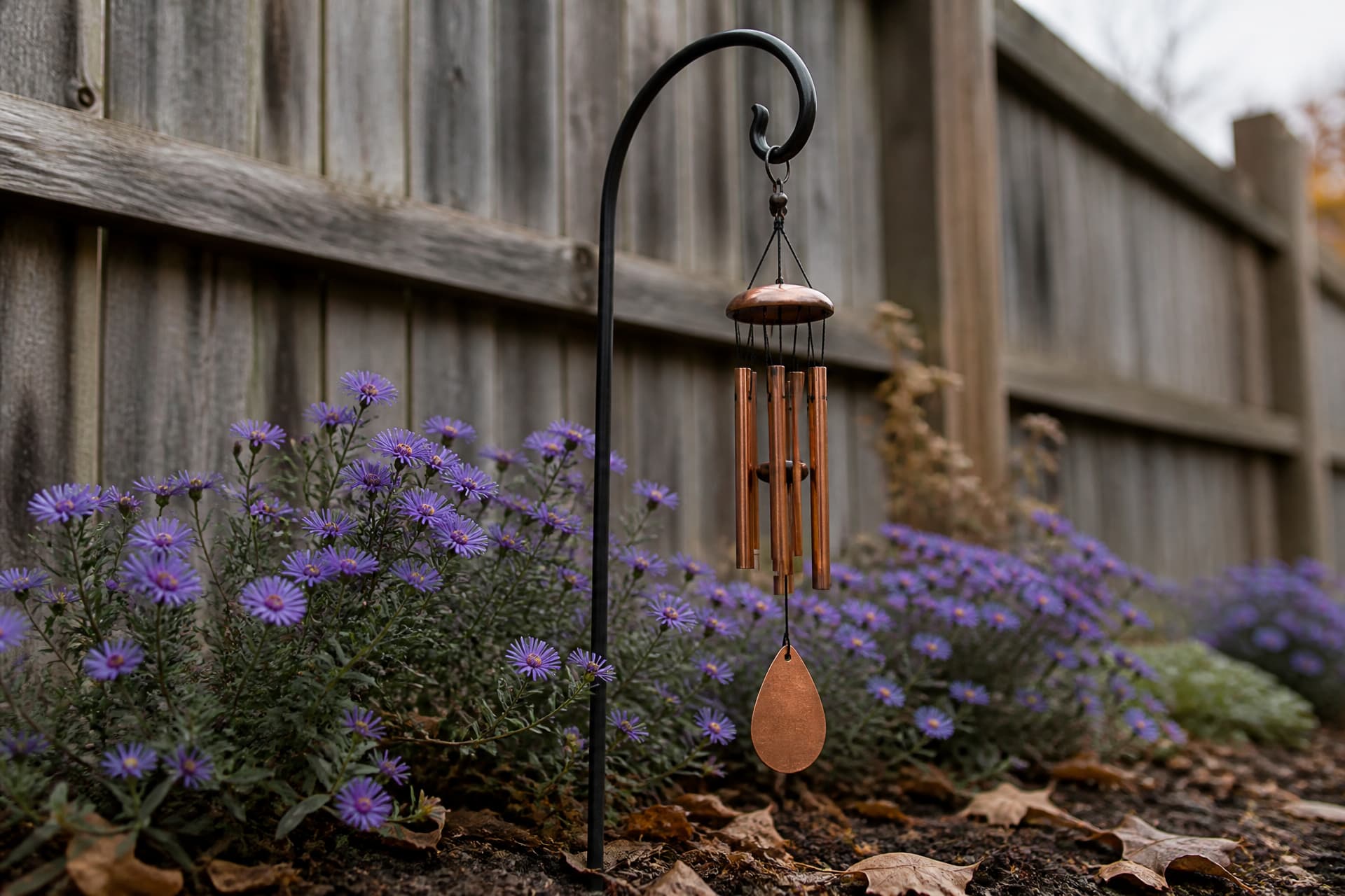 A shepherd's hook set into a flower bed of low purple asters with a small copper wind chime hanging from it, a weathered timber fence behind, overcast afternoon light