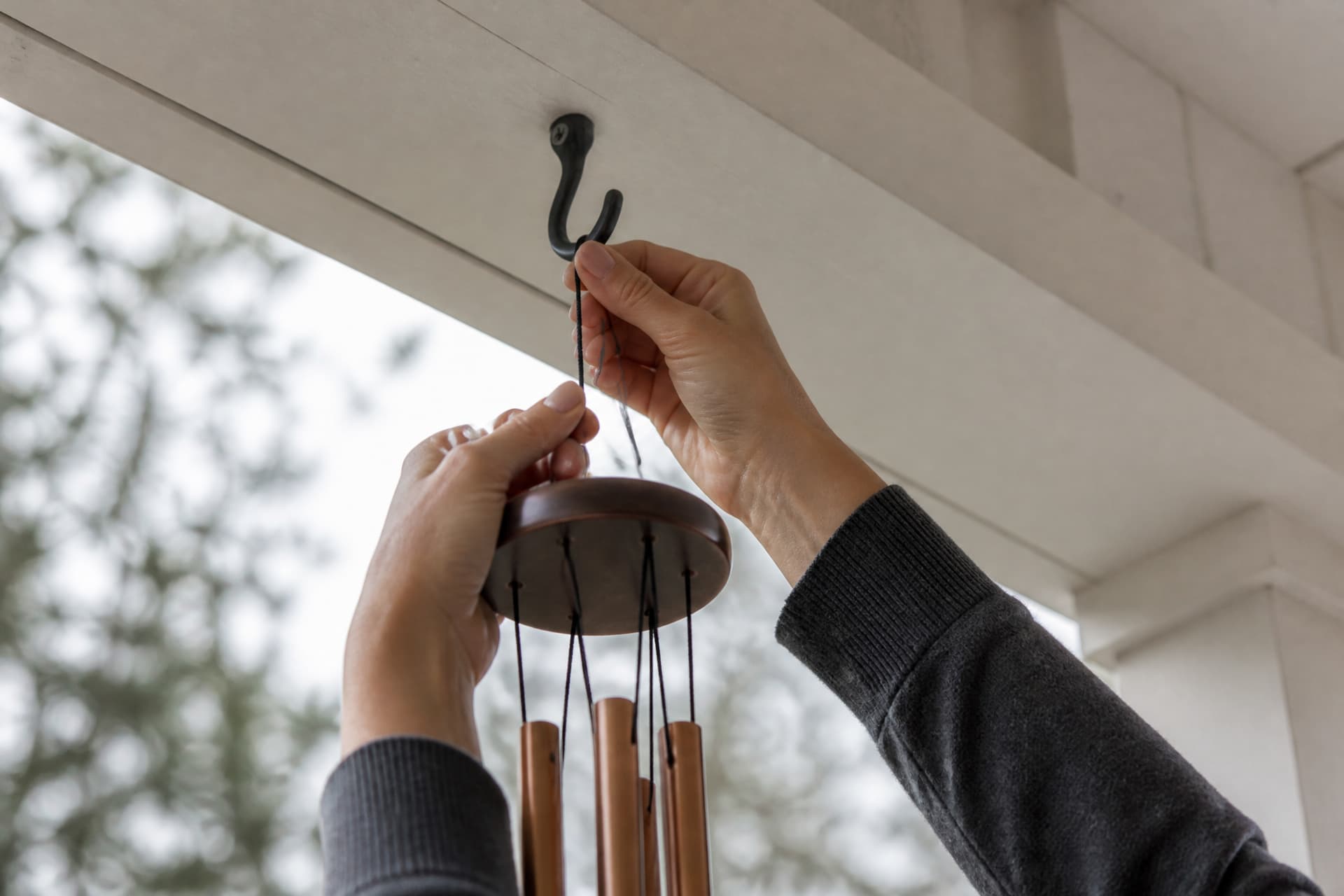 A woman's hands reaching up to settle the cord of a copper wind chime onto a black iron hook screwed into the white painted underside of a porch eave, her charcoal cardigan sleeves falling back at the wrist, the tubes hanging still below